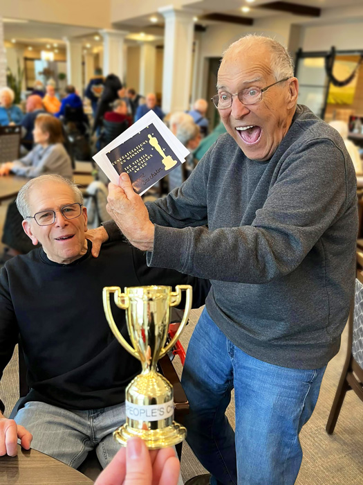 Two senior residents excitedly celebrating with a small trophy and a certificate in a lively senior living community setting, highlighting the joy of winning and friendly competition.