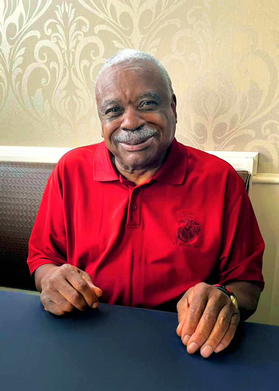 Smiling senior resident in a red polo shirt with a U.S. Marine Corps logo, seated at a table against an elegant patterned wallpaper, radiating warmth and pride in community living.