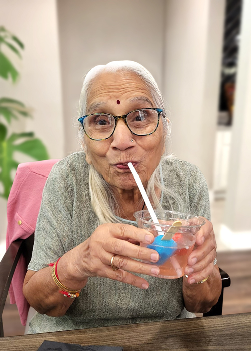 Joyful senior resident with a bindi, sipping a colorful drink through a straw, capturing a moment of delight and celebration in a vibrant community setting.