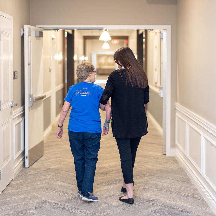 Back view of a senior resident in a blue shirt walking down a hallway with a young woman in black, holding onto her arm for support, as they share a moment of companionship.