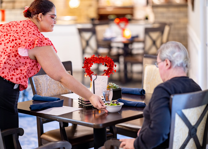 Caregiver in a red blouse serves a salad to a senior senior resident seated at a beautifully set table in a cozy dining area, highlighting attentive service and community dining.