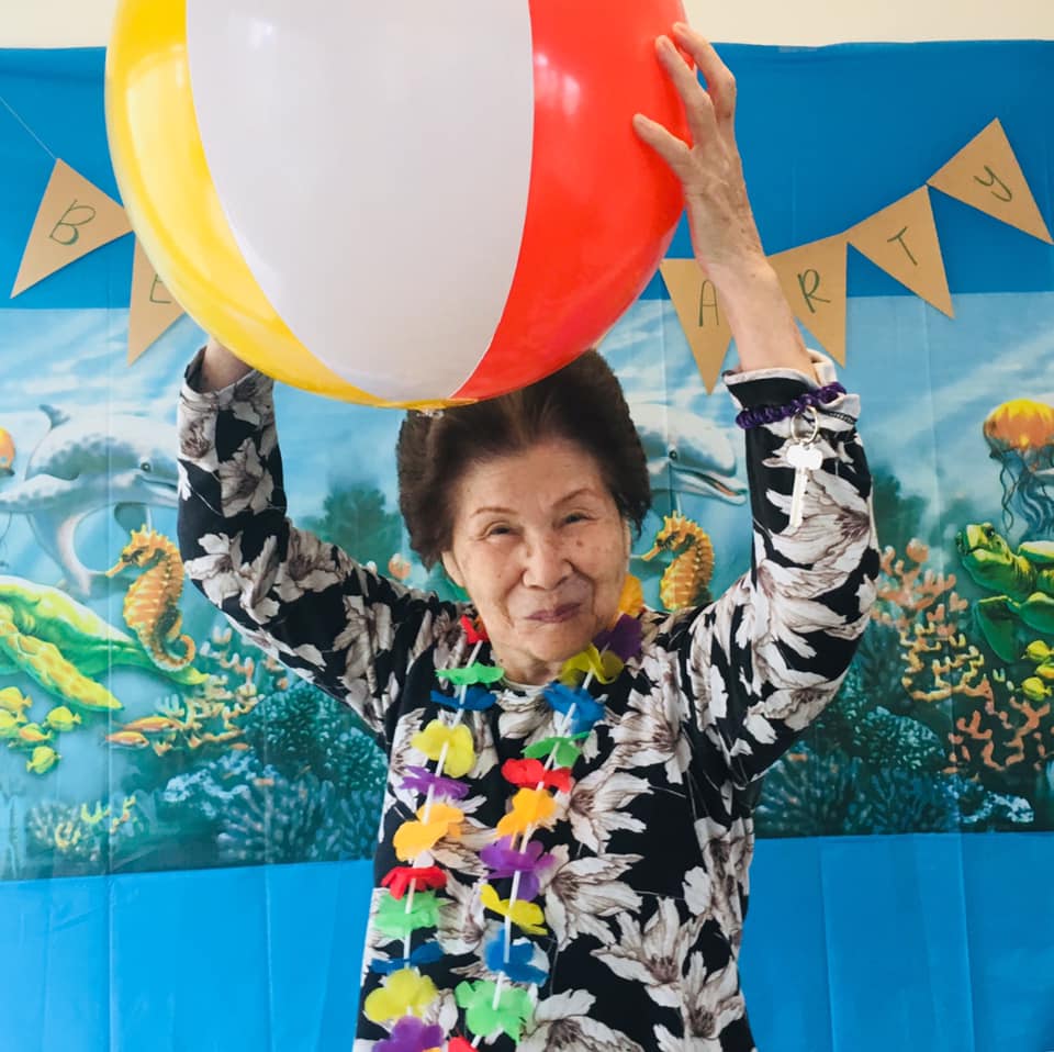 Senior resident with a joyful smile holds a colorful beach ball above her head, wearing a floral shirt and a bright lei, celebrating a summer-themed event with enthusiasm.