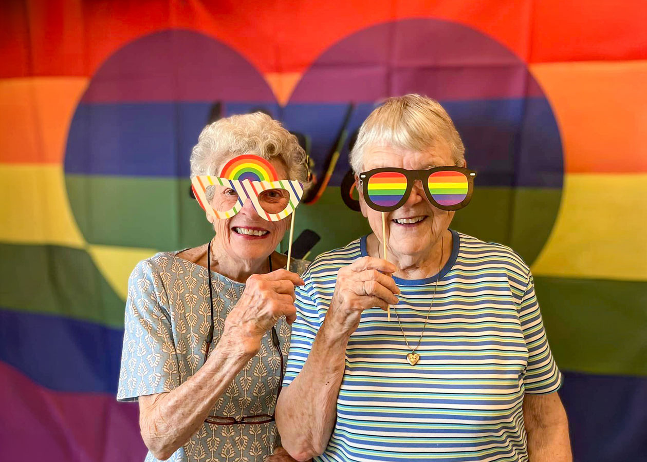 Two senior residents celebrate Pride Week by posing with rainbow-themed props in front of a colorful Pride flag backdrop, smiling and showing their support for LGBTQ+ pride.