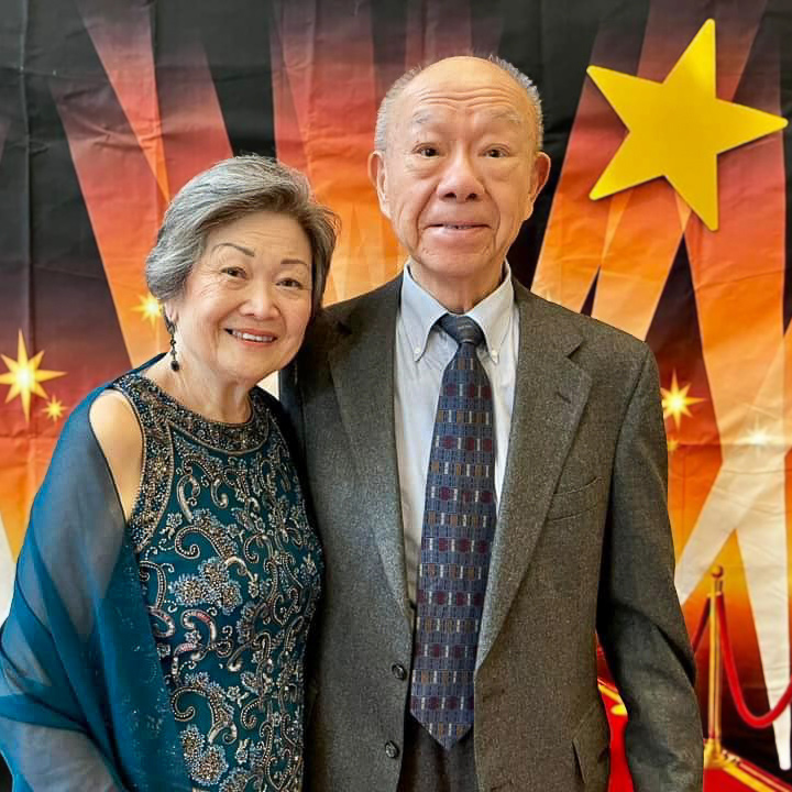 Elegant senior resident couple posing together at an event, with the woman in a detailed blue gown and the man in a gray suit, standing in front of a red carpet-themed backdrop.