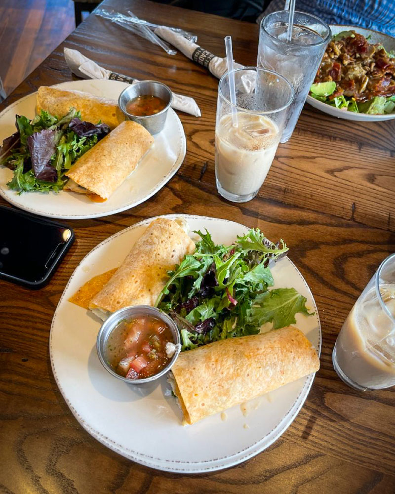 Two plates of fresh wraps served with mixed greens and salsa, alongside iced coffee and water glasses, on a wooden table at a cozy restaurant setting.