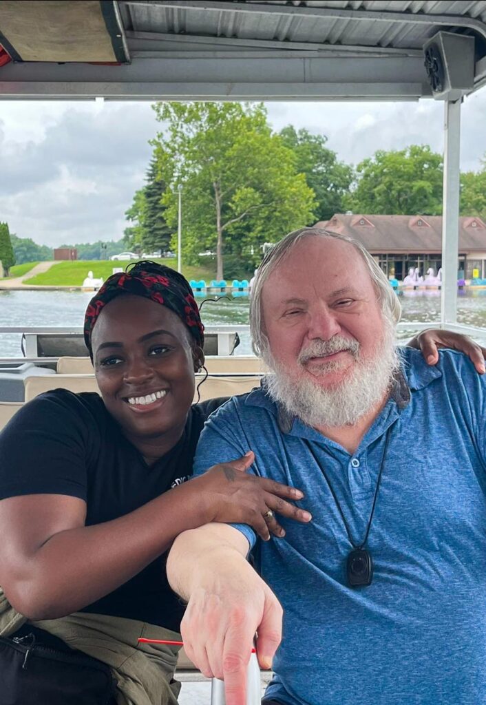 Employee and senior resident smiling and sitting side by side on a boat, with a scenic view of a lake and green trees in the background at The Township Senior Living.