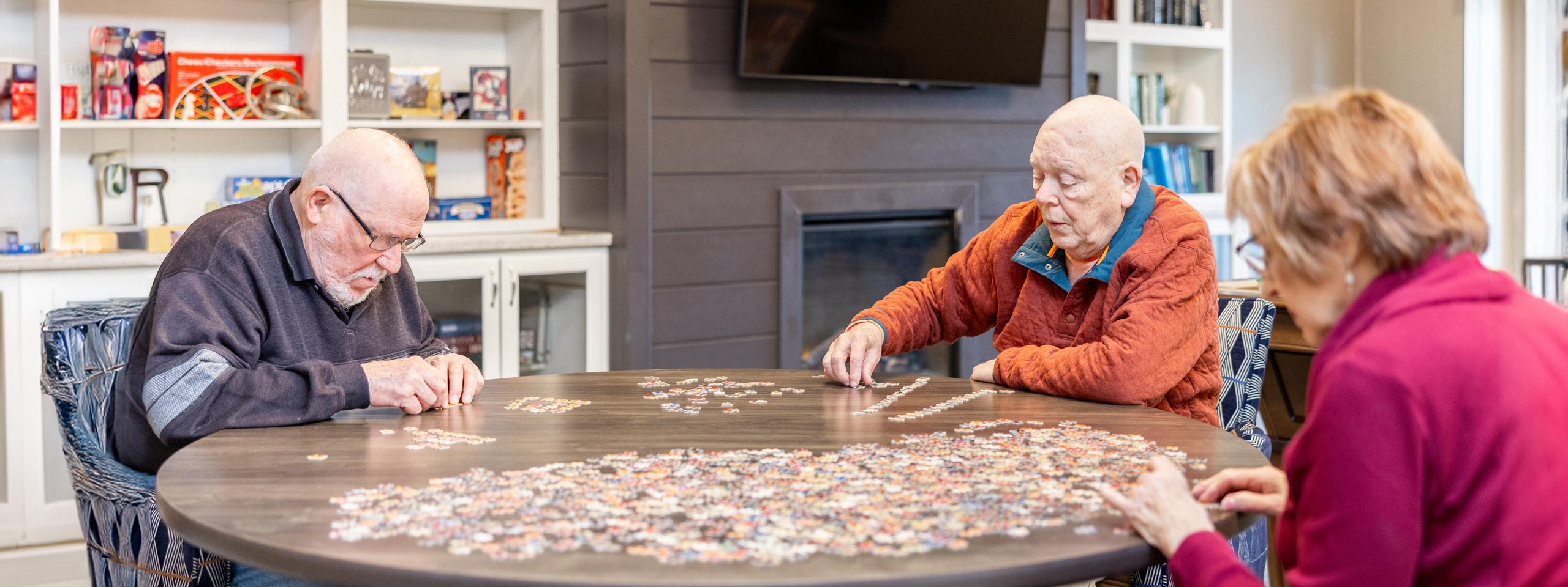 Three seniors focused on assembling a large puzzle together on a round table in a cozy community room, engaging in a stimulating and social activity for mental wellness.