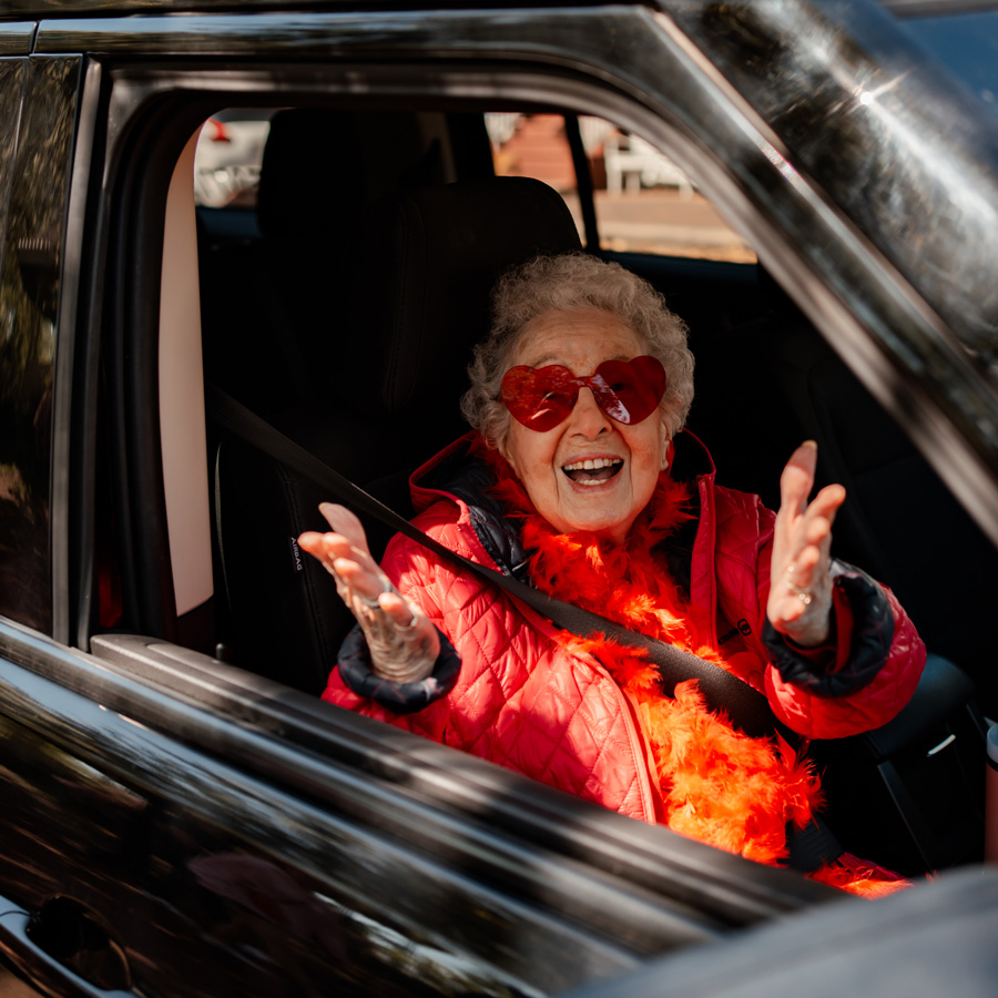 Senior resident in a car, joyfully wearing red heart-shaped sunglasses and a matching feather boa, enthusiastically waves while dressed in a bright red jacket.