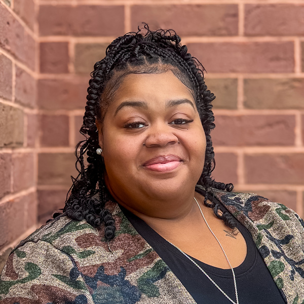 Beverly Merriweather, Business Office Director at University Senior Living, smiling in front of a red brick wall while wearing a camo-patterned cardigan and necklace.