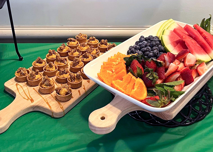 Colorful fruit platter with watermelon, strawberries, blueberries, oranges, and melon slices next to mini pecan pie tarts drizzled with caramel on a cutting board.