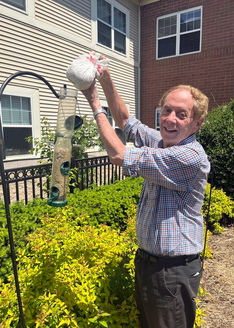 Senior resident smiling while refilling a bird feeder on a sunny day, surrounded by green shrubs and a brick senior living building in the background.