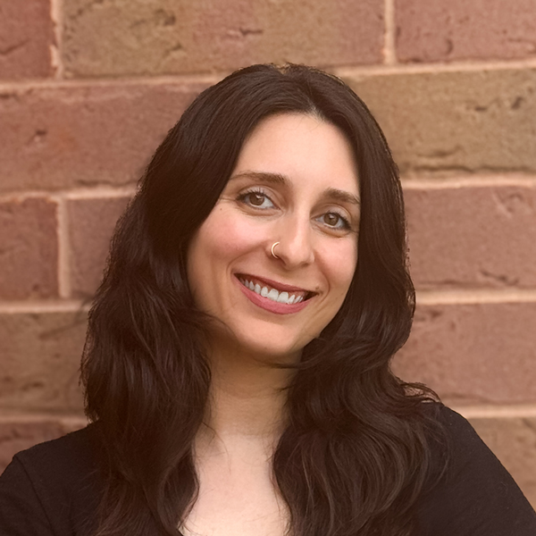 Glenna Benitez, Senior Living Director at University Senior Living, smiling with long dark hair and a nose ring, standing in front of a brick wall.