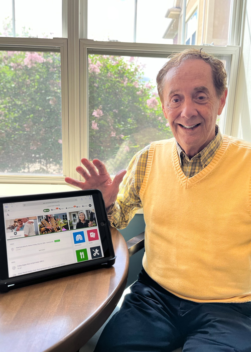Senior man sitting by a window and smiling while showcasing a tablet displaying the Arrow app on a table in front of him.