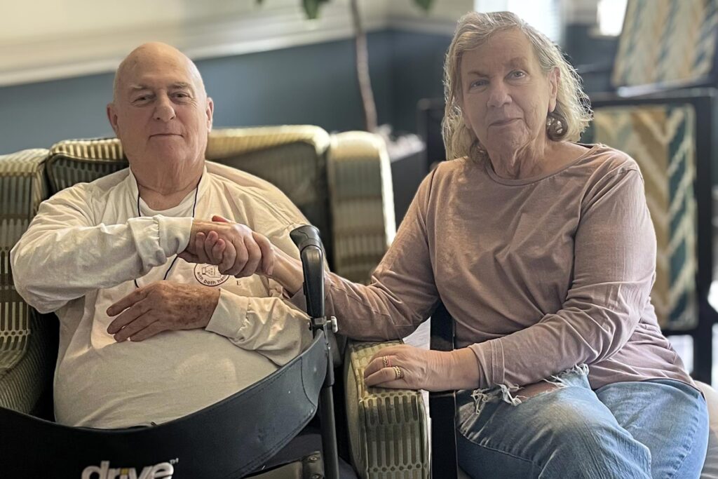 Senior couple sitting side by side in a cozy community lounge, holding hands warmly and sharing a quiet, tender moment together in a supportive environment.