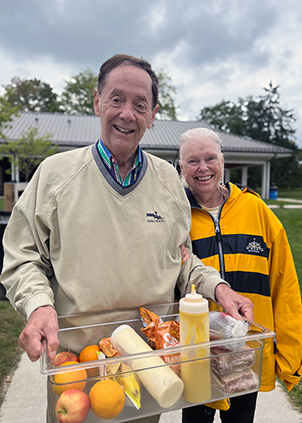 A senior couple smiles outdoors, the man holding a container full of fruits, sauces and meal ingredients.