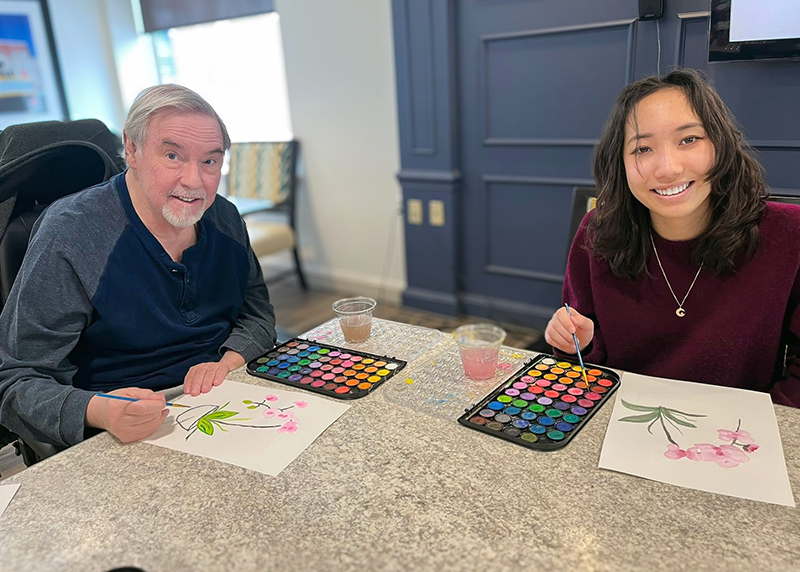 A senior man and loved one smile brightly during a relaxed art session at University Senior Living.