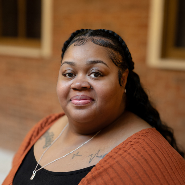 Beverly Merriweather, Business Operations Director at University Senior Living, looking at the camera in a professional outdoor headshot with braided hair pulled back, a rust-colored cardigan, layered necklaces, and a softly blurred brick background.