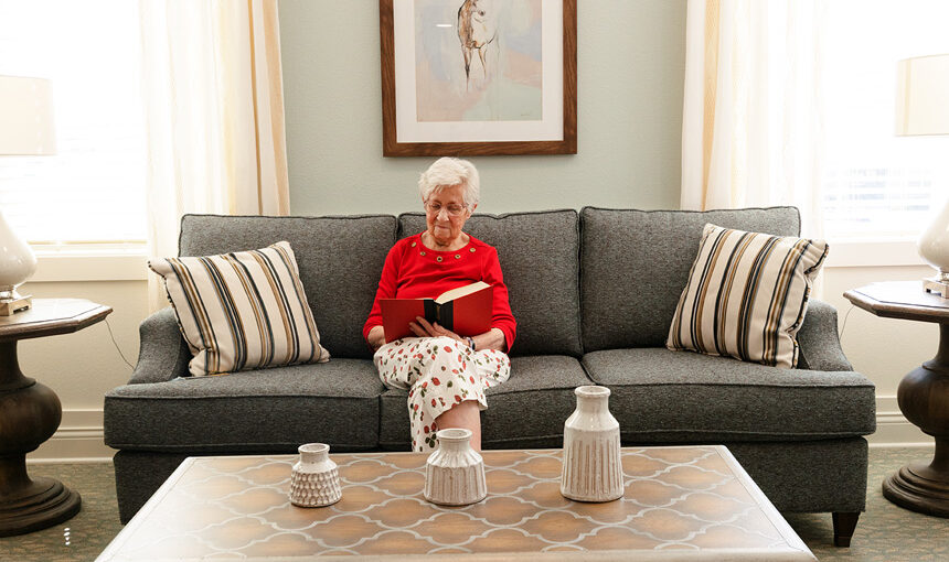 Resident reads a book on a gray sofa in a bright, comfortable lounge at The Madison Senior Living.