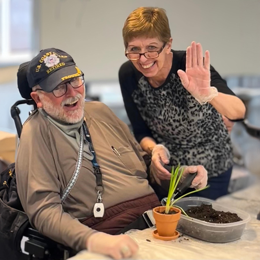 A senior man in a wheelchair smiles as a woman holds a plant, celebrating National Gardening Month together.