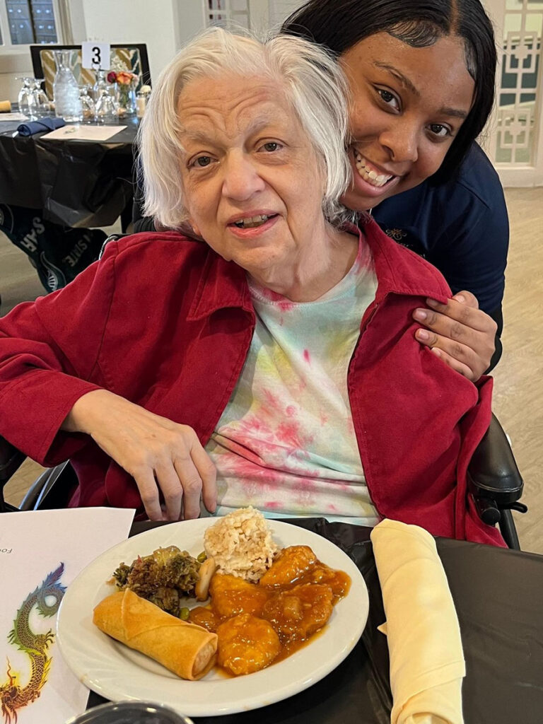 Employee and senior resident share a warm hug at a table with food, captured in a joyful photo - touching moment between generations.