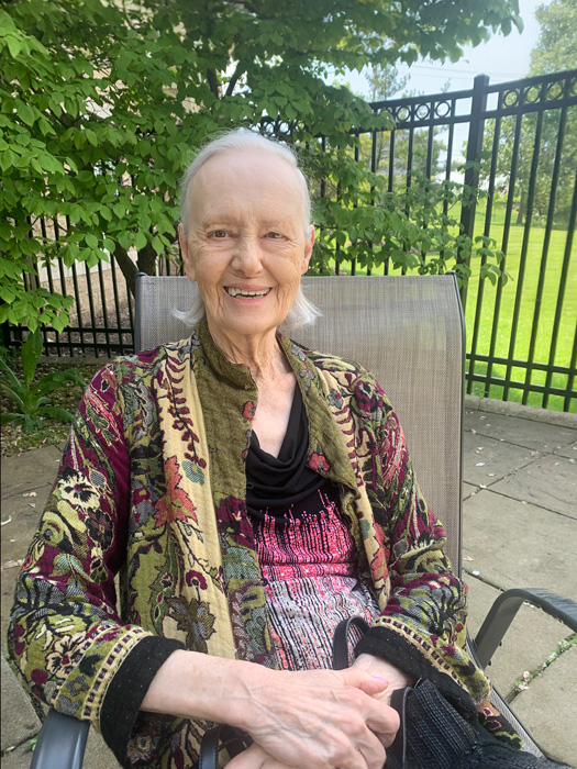 Senior woman with gray hair, wearing a colorful patterned jacket, smiling while seated outdoors in a garden area with a black fence in the background.