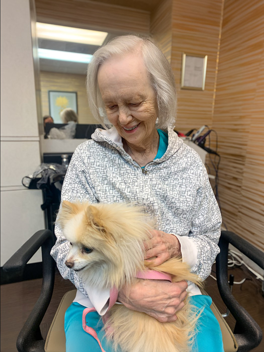 Senior woman with gray hair, wearing a patterned hoodie and blue pants, smiling while holding a small fluffy dog in her lap indoors.