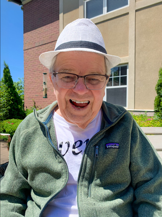 Senior man wearing a white hat, glasses, and a green jacket, smiling broadly while sitting outdoors in a garden area on a sunny day.