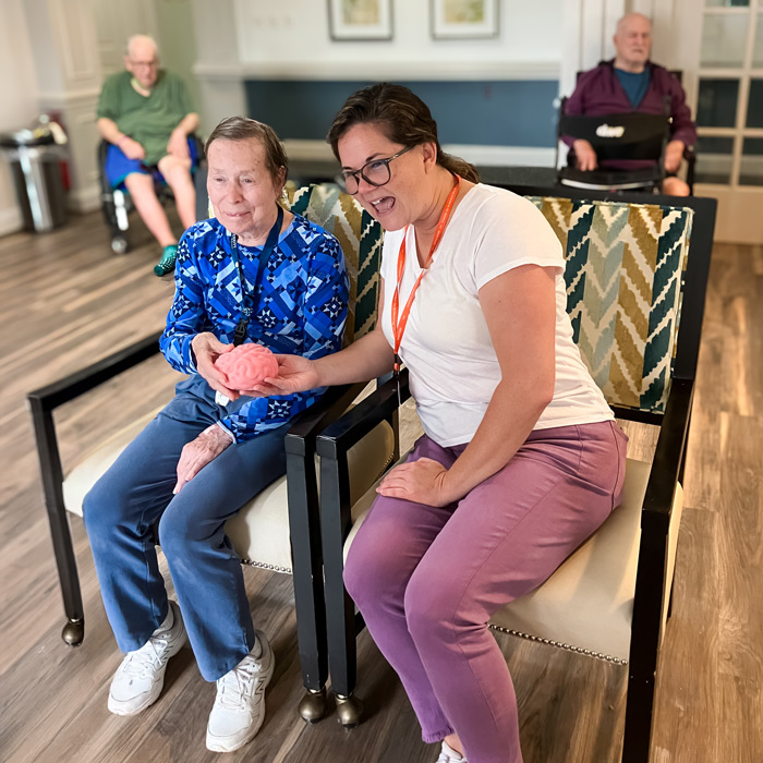 A senior resident and a staff member share a joyful moment while holding a pink brain model, engaging in a fun and educational activity in a bright, comfortable room.