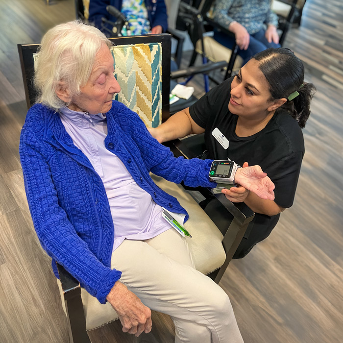 A senior resident has her blood pressure checked by a staff member during 'Wellness Wednesday,' both engaging warmly in a comfortable room.