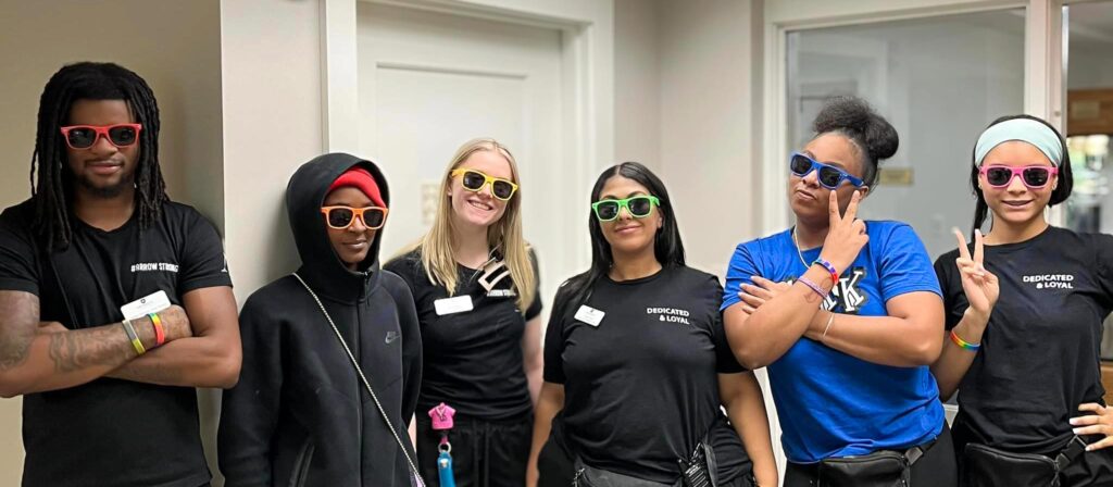 Six team members wearing colorful sunglasses and posing playfully during a fun event at a senior living community, showcasing their 'Dedicated & Loyal' spirit.