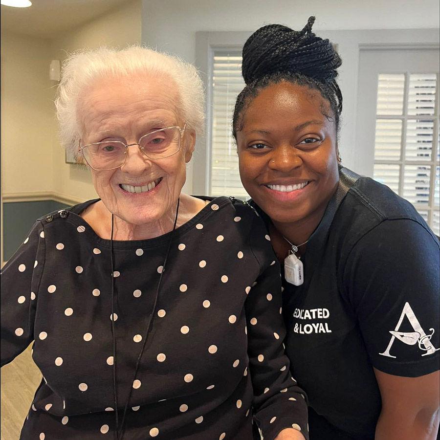 Elderly resident with white hair and glasses smiles warmly alongside a cheerful caregiver, both enjoying a moment together at a senior living community.