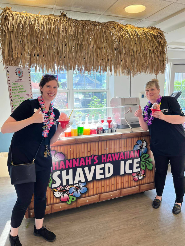 Two women in black shirts and leis smile and give thumbs up while enjoying shaved ice from a colorful 'Hannah's Hawaiian Shaved Ice' stand.