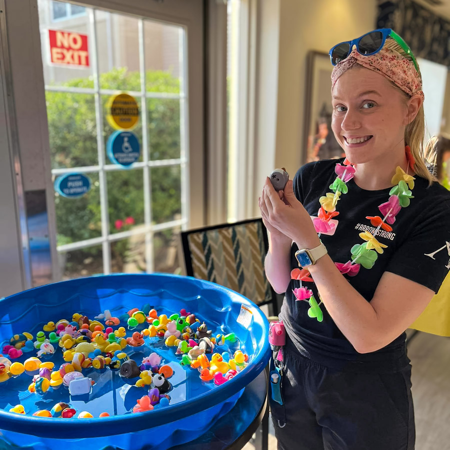 A woman in a black shirt, colorful lei, and sunglasses smiles while holding a rubber duck near a kiddie pool filled with various rubber ducks at a summer-themed event.