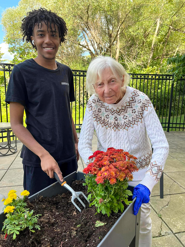 A senior resident and a young staff member enjoy gardening together outdoors. The resident, wearing a sweater, is tending to flowers with a small gardening tool.