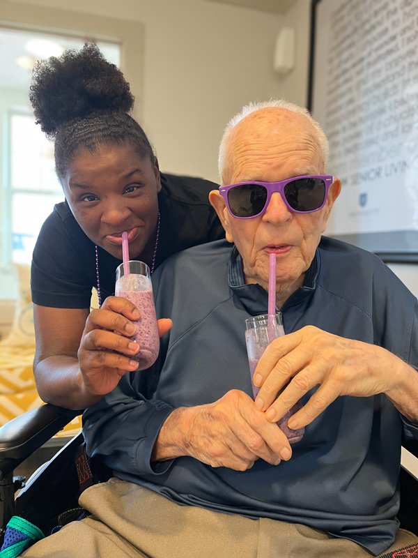 A senior resident and a staff member sip on smoothies together. The resident, wearing sunglasses, smiles while enjoying the moment with the staff member.