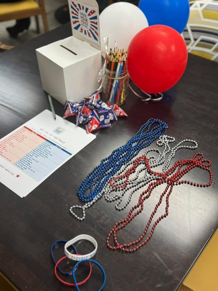 A table decorated with red, white, and blue balloons, patriotic beads, a voting box, and themed treats, set up for a senior living community voting event.