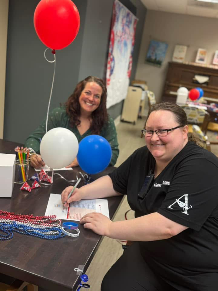 Two senior living employees smile while participating in a voting-themed event, surrounded by patriotic decorations, a ballot box, and celebration materials.