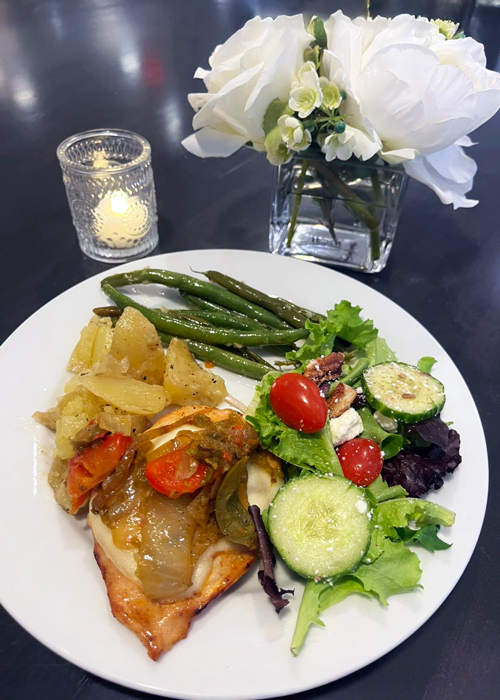 Plate of smothered chicken, herb-crusted potatoes, green beans, and salad beside a lit candle and flower vase during Earth Month farm-to-table dinner.