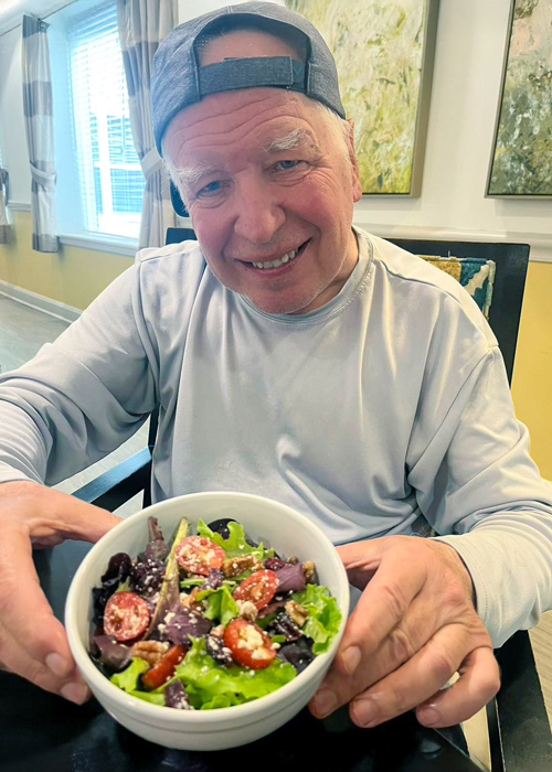 Smiling senior resident holds a fresh garden salad with tomatoes, pecans, and feta cheese during a candlelight Earth Month dinner celebration.
