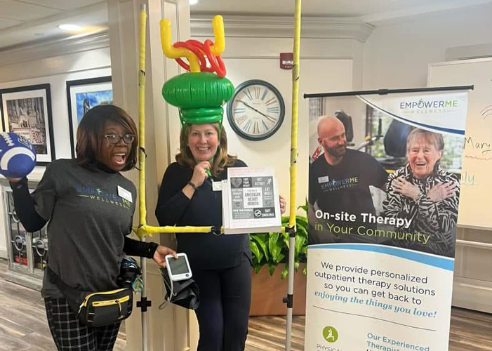 Two women dressed for a fun therapy event, one holding a football and the other wearing an inflatable goalpost hat, standing beside a therapy services banner.