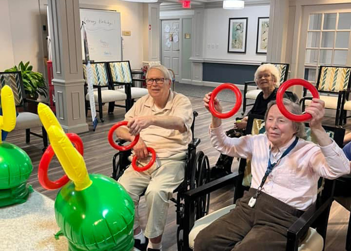 Senior residents seated at a table enthusiastically playing a ring toss game with inflatable goalpost hats during a group activity indoors.