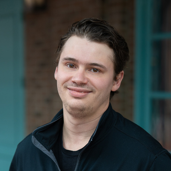 Christopher “Chris” King, Culinary Director at University Senior Living, smiling in a professional headshot, wearing a black zip-up jacket, with a softly blurred brick and window background.