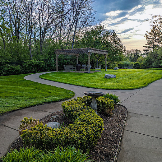 Walking paths outside University Senior Living, landscaped with mulch beds, foliage and trees, and featuring a large covered seating area.